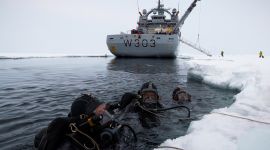 Tre dykkere i havet, ved iskanten. I bakgrunnen ser vi kystvaktens isbryter. 