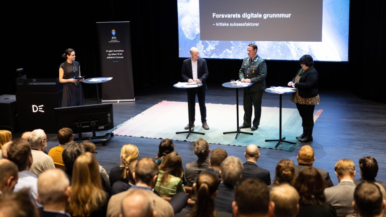 paneldeltakere står på scenen foran en power point i forgrunnen skimter vi hodene til de i salen