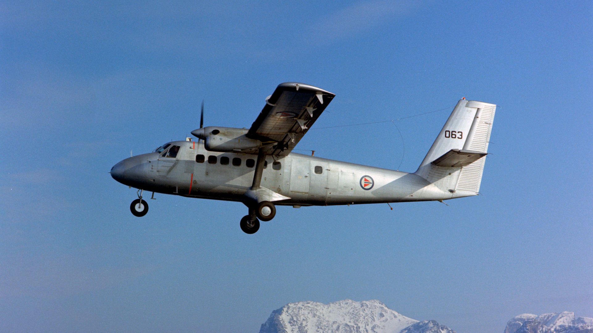 En sølvfarget Dehavilland Twin Otter i Nord Norge 1978 flyr med fjell i bakgrunnen.