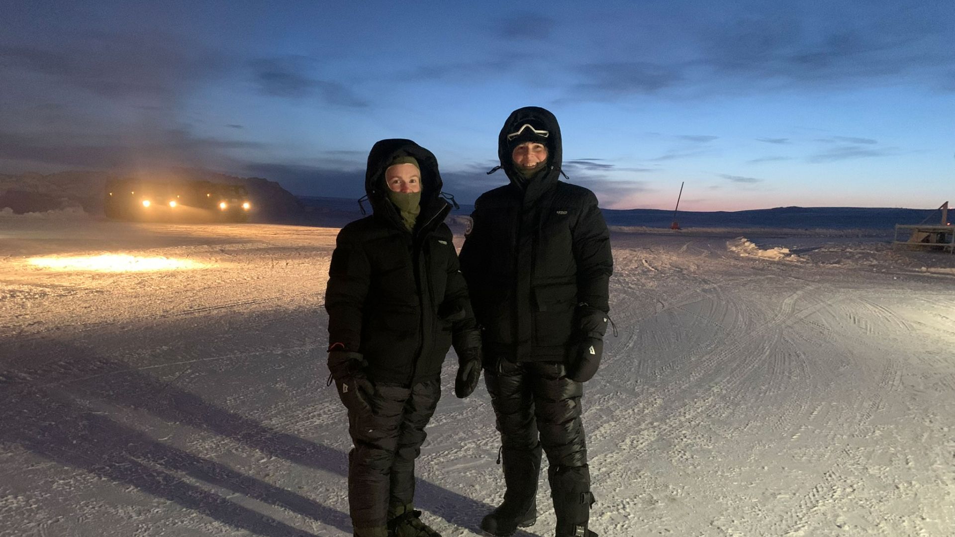 Julie Renberg og Hilde Teien (th) i mørke vinterklær i et snødekt landskap. To biler med lys på i bakgrunnen.