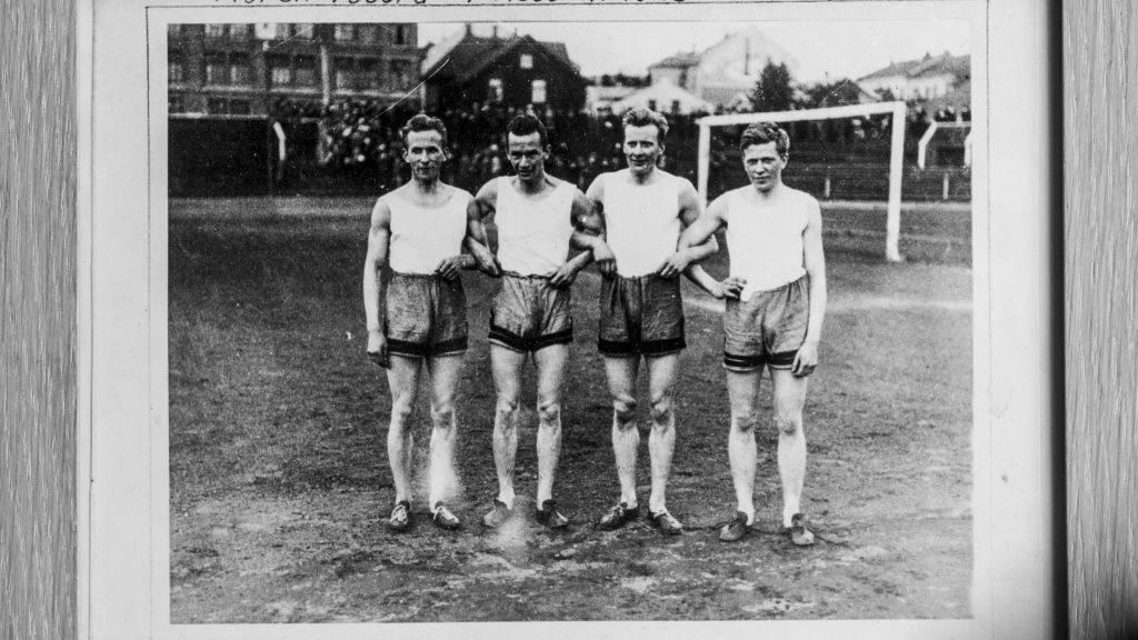 Gruppebilde av fire menn i shorts og T-skjorter. Bildet er gammel og i ramme med en tekst nederst i bildet.