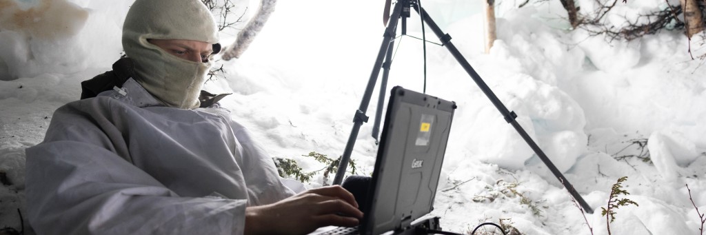 Soldat sitter i snøhule med en liten datamskin foran seg. Han har buff foran ansiktet. I åpningen står en kikkert på stativ.