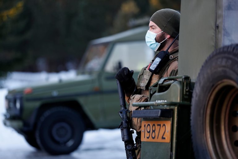 Vaktsoldat med munnbind og våpen. Står mellom to Mercedes feltvogner.