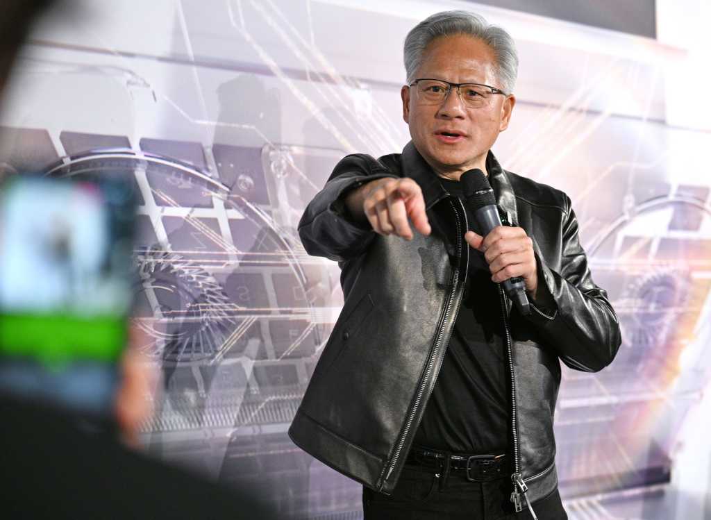 Jensen Huang i svart skinnjakke. Den ene hånda peker mot fotografen. I den andre holder han en mikrofon.