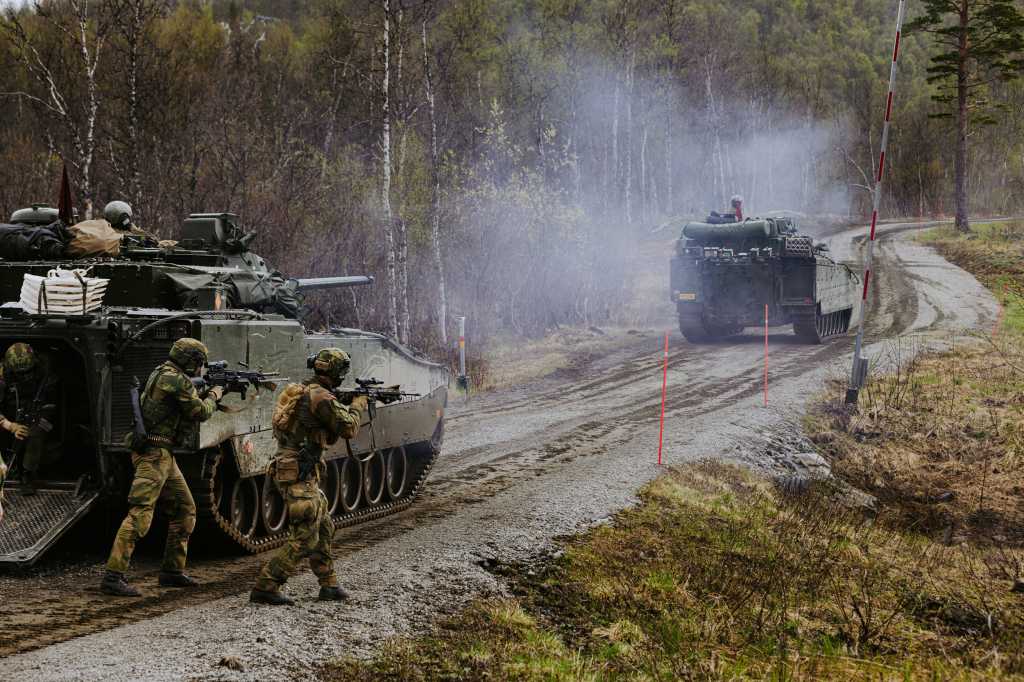 To CV90 på en grusvei med en oppslått bom og brøytestikker langs veikanten. To soldater med gevær går til høyre for den bakerste kampvognen.