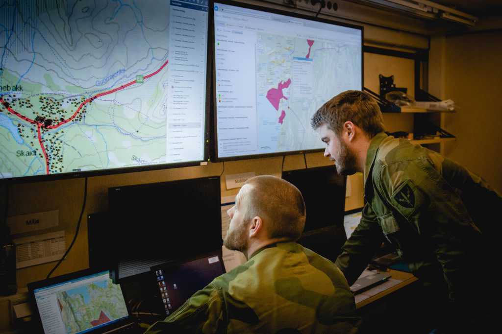 To menn i militæruniform sitter og ser på skjermer med militære kart og informasjon