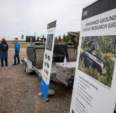 Rollups oppstilt foran militært materiell utendørs. Tre mennesker står  i bakgrunnen. Foto