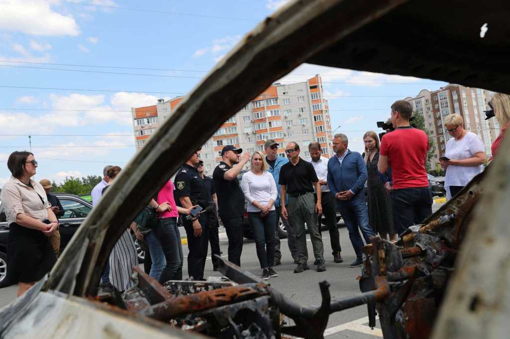 En rekke personer står foran et ødelagt kjøretøy. En politimann snakker til forsamlingen.