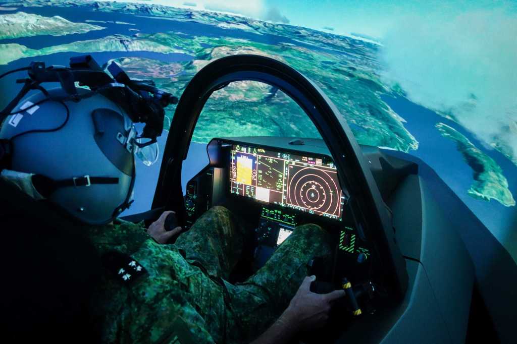 En person med hjelm sitter i en F-35 simulator. Vi ser instrumentpanelet og et landskap i bakgrunnen.