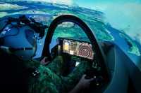 En person med hjelm sitter i en F-35 simulator. Vi ser instrumentpanelet og et landskap i bakgrunnen.