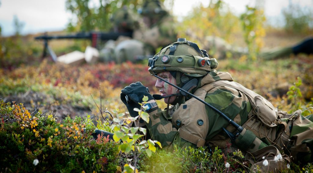 soldat i kamuflasje ligger i norsk lyng og speider med kikkert