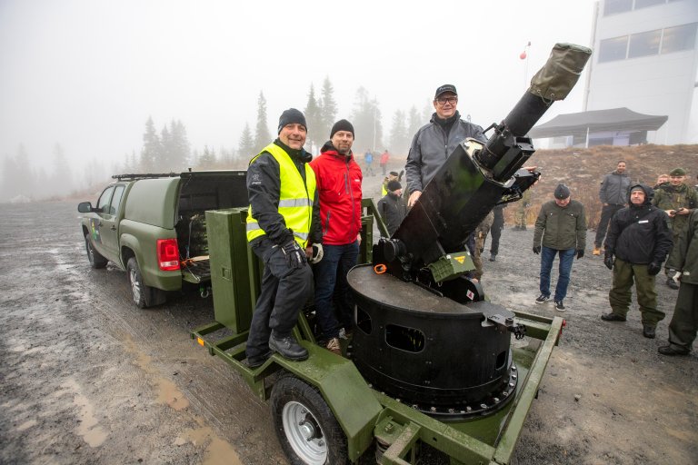 Tre menn står ved en bombekaster på en tilhenger ute i skogen