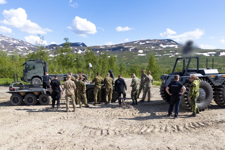 En gruppe soldater foran fire ubemannede kjøretøyer. Fjell, sol og blå himmel.