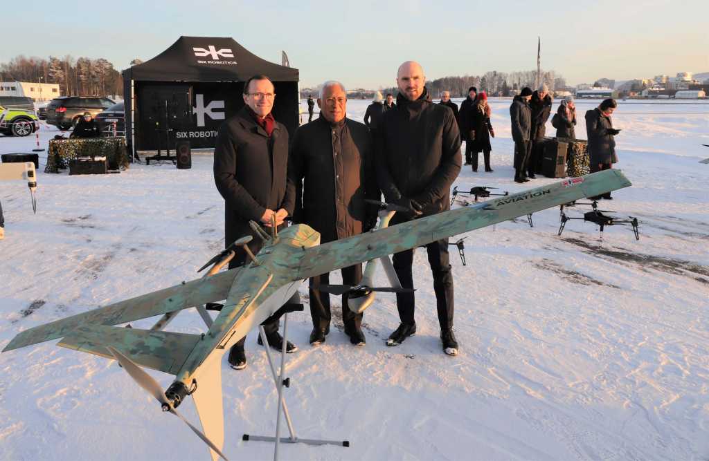 Tre menn poserer bak en drone på en snølagt flyplass.