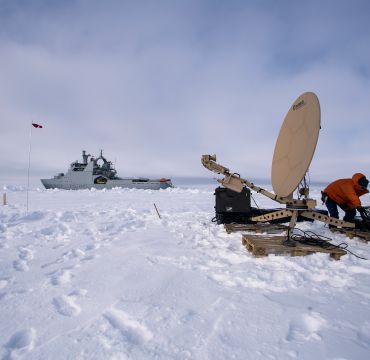 en fregatt ligger i isen. i forgrunnen ser man tre forskere i boblejakkre som ordner med en parabolantenne
