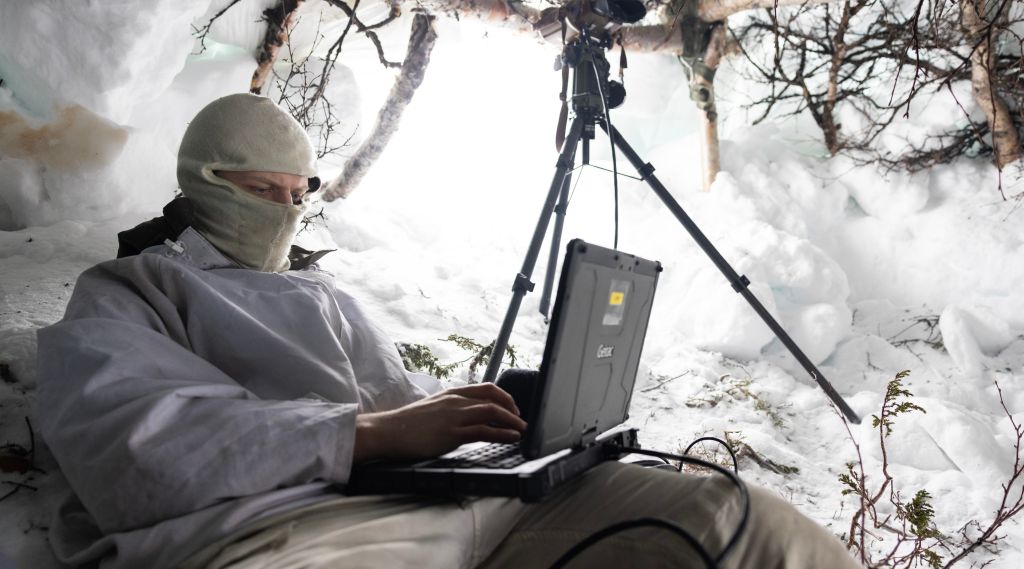 Soldat sitter i snøhule med en liten datamskin foran seg. Han har buff foran ansiktet. I åpningen står en kikkert på stativ.