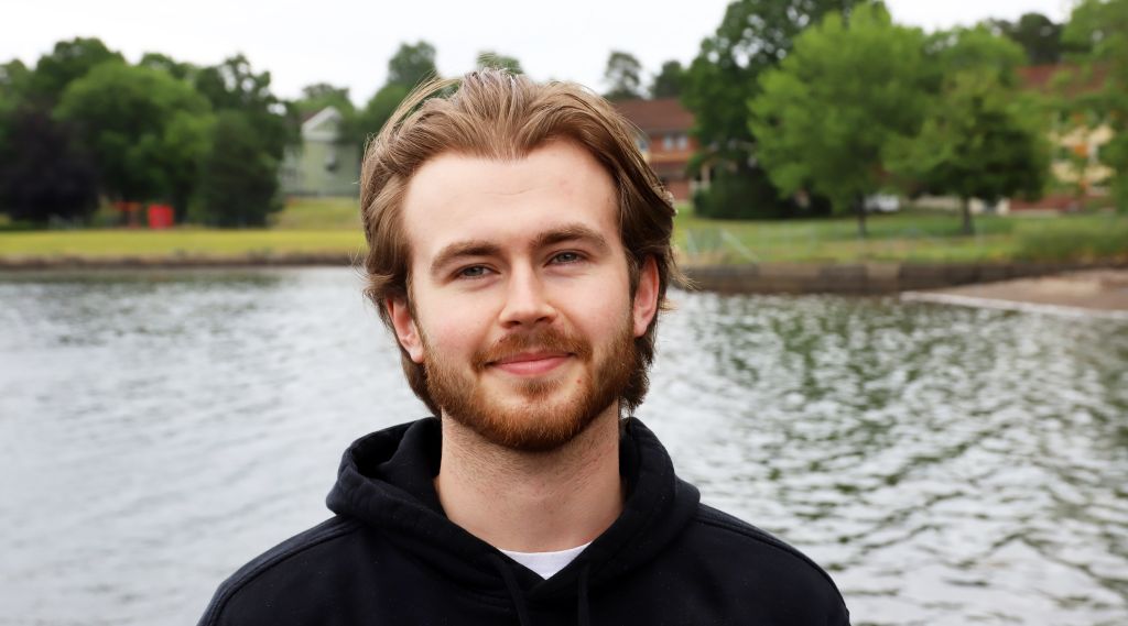 Ung mann i hettegenser står utendørs med havet i bakgrunnen. Ser rett i kamera og smiler