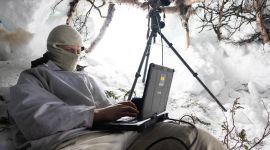 Soldat sitter i snøhule med en liten datamskin foran seg. Han har buff foran ansiktet. I åpningen står en kikkert på stativ.