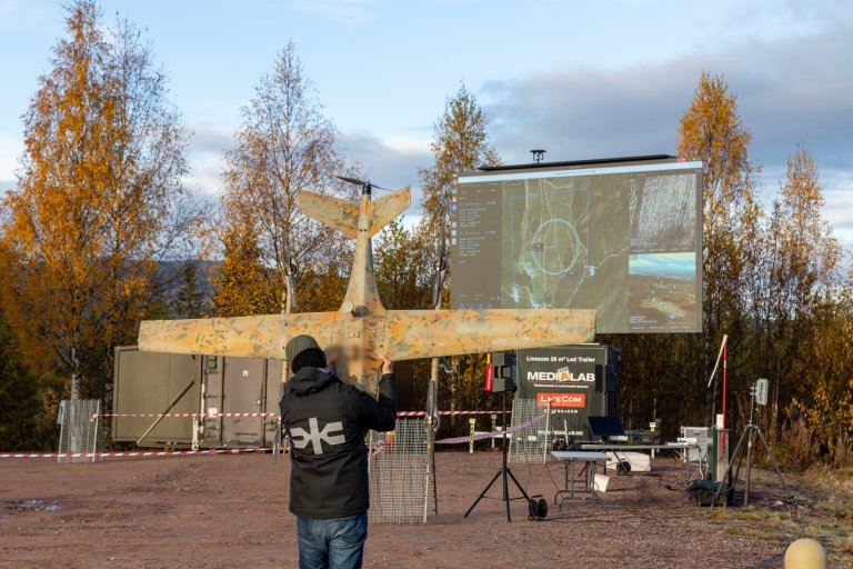 En mann bærer en drone. I bakgrunnen ser vi en stor skjerm og noen utstillingsbord. Utendørs. Høst.