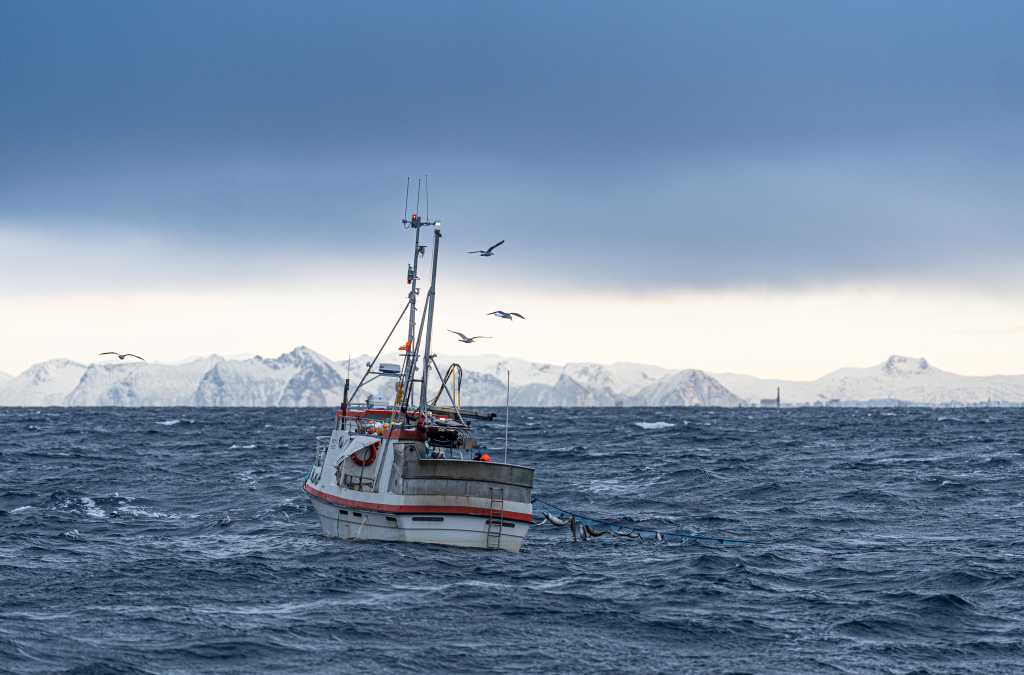 Bilde av fiskeskøyte på havet et sted i Nord Norge. Snødekte fjell i bakgrunnen.