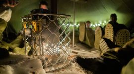 Vi er inne i et telt. Det er nærbilde av en ovn og flere soldatføtter som varmer seg på den