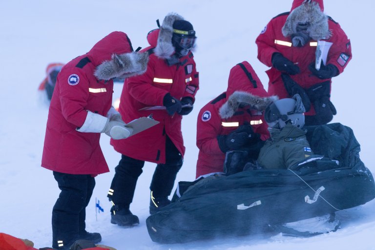 Fire forskere i røde jakker med hette og pelskrage står bøyd over en person i en pulk. Forskerne har spørrskjemaer i hendene. Det blåser kraftig.