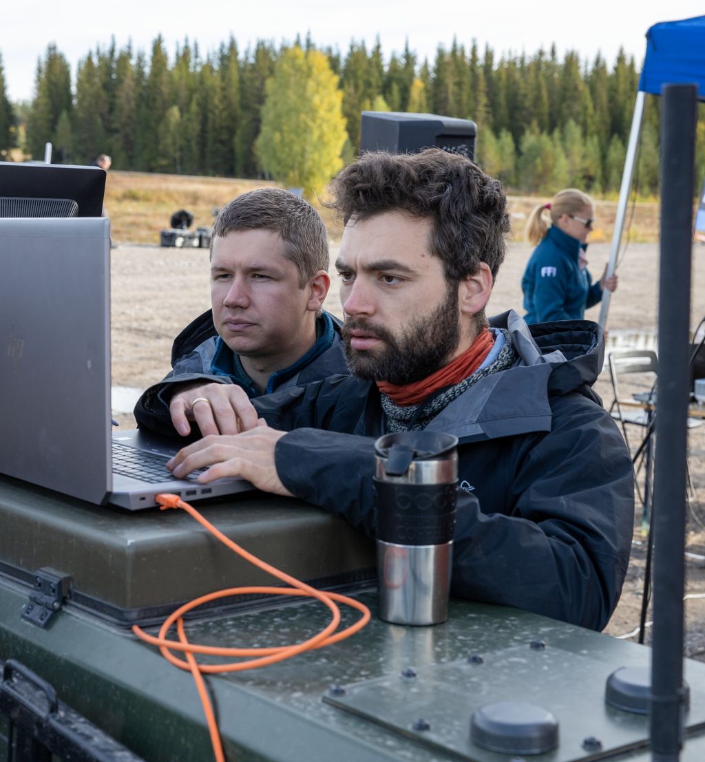 To menn ser på laptop mens de står utendørs på et militært øvingsfelt