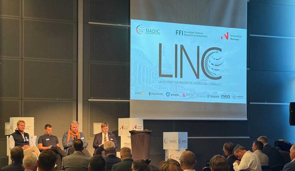 Fire menn sitter på en scene. Logoen til LINC vises på en stor skjerm.