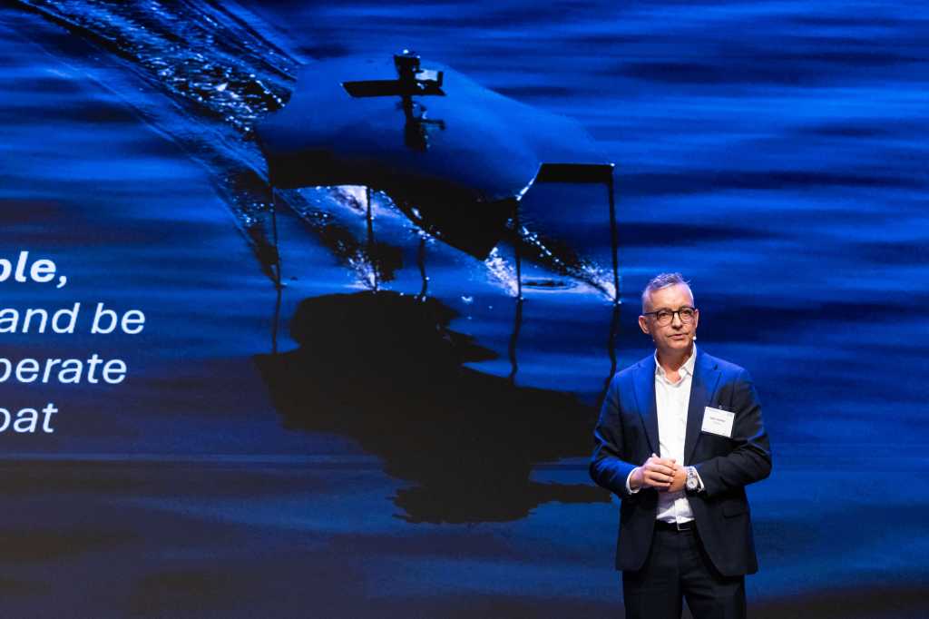 Mann i dress foran bildet av en sjøgående drone.