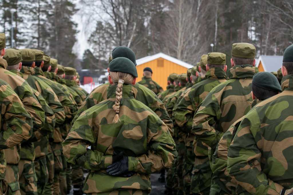 Soldater på oppstilling i grønn feltuniform. Alle står med ryggen til. I midten en kvinne med en lang flette.