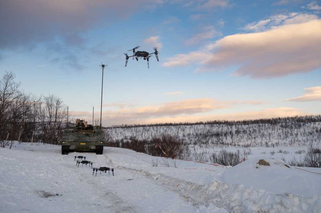 En drone flyr i luften. Tre andre står på bakken klare til takeoff foran en stridsvogn. Vinter. Nordnorsk landskap.