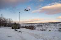 En drone flyr i luften. Tre andre står på bakken klare til takeoff foran en stridsvogn. Vinter. Nordnorsk landskap.