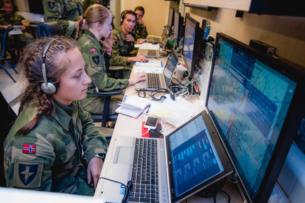 en ung kvinne i grønn militæruniform og norsk flagg på armen sitter med hodetelefoner og ser på flere dataskjermer