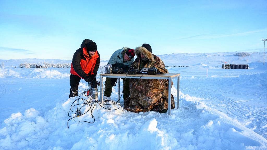 Tre menn står ved et skrivebord ute i snøen og studerer instrumenter/ledninger