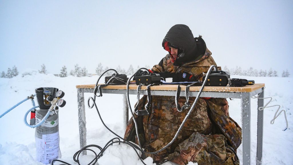 Mann som sitter godt innpakket ved et skrivebord ute i snøen. På bordet er det masse utstyr og ledninger