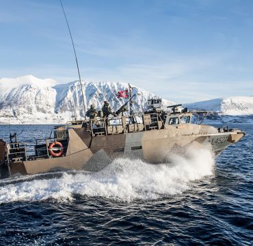 En Forsvars-båt kjører i fyll farg med snødekte fjell i bakgrunnen og klar, blå himmel
