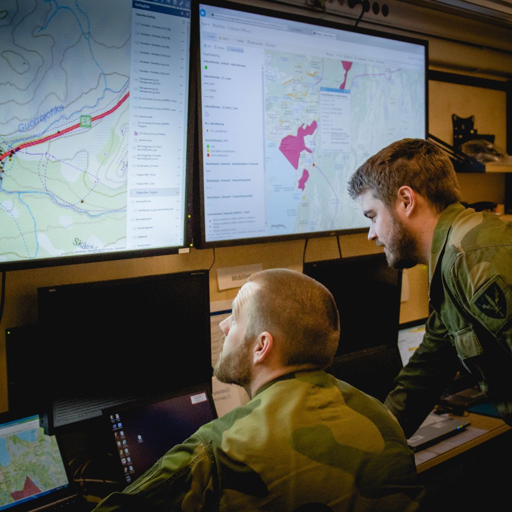To menn i militæruniform sitter og ser på skjermer med militære kart og informasjon