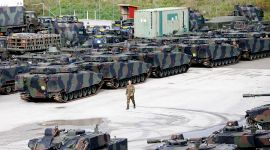 et oversiktsbilde viser en stor plass med mange stridsvogner og andre militære kjøretøy som er parkert. Midt på plassen går en mann i hæruniform