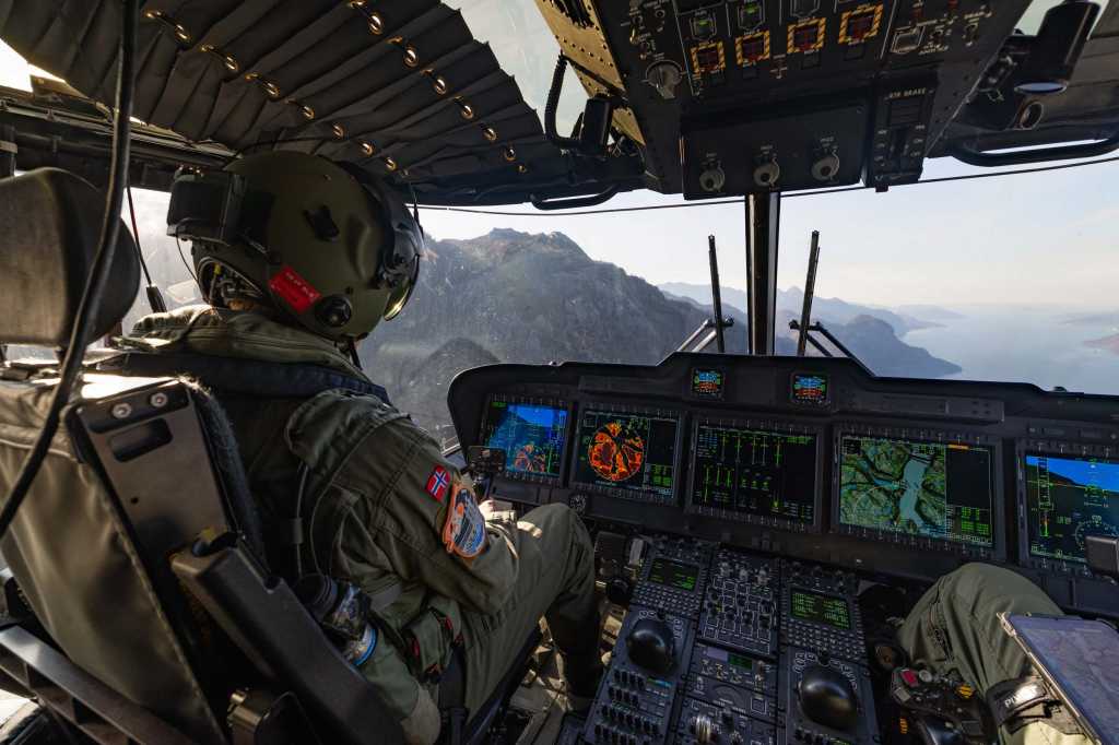 Utsikt fra cockpit til et helikopter. Fleire instrument lyser opp. Vi ser ryggen og hjelmen til piloten.