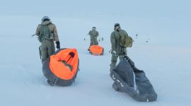 Tre piloter med flydress og helm går over en slette dekket av is og snø. De trekker på hver sin oppblåsbare flåte med overlevelsesutstyr.