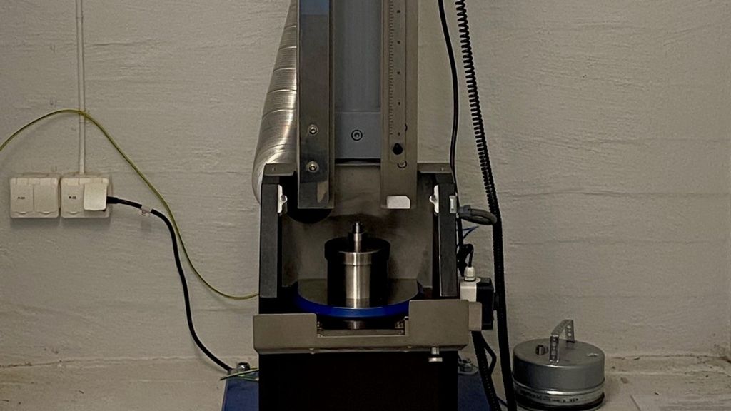 Maskin som sender vekter ned mot en metallbeholder. Ved siden av maskinen ligger et gult måleinstrument.
