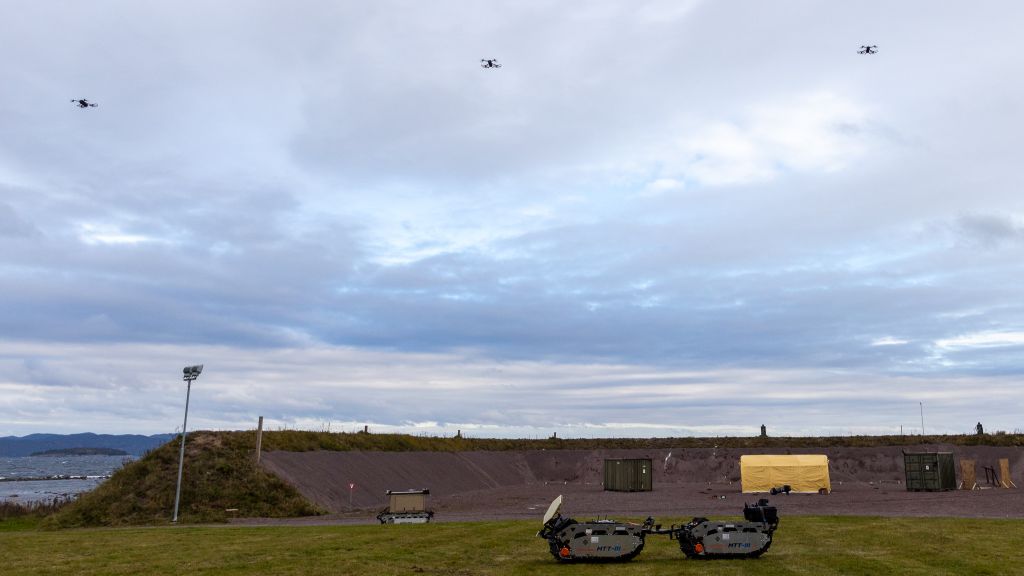 Tre overvåkingsdroner svever i lufta. På bakken i forgrunnen en ubemannet beltevogn.