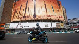 Mann på motorsykkel kjører forbi en gigantisk plakat av missiler midt i teheran. Blå himmel. Sommer. Trafikk.