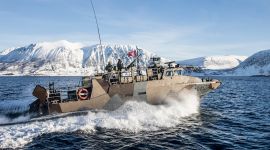 En Forsvars-båt kjører i fyll farg med snødekte fjell i bakgrunnen og klar, blå himmel