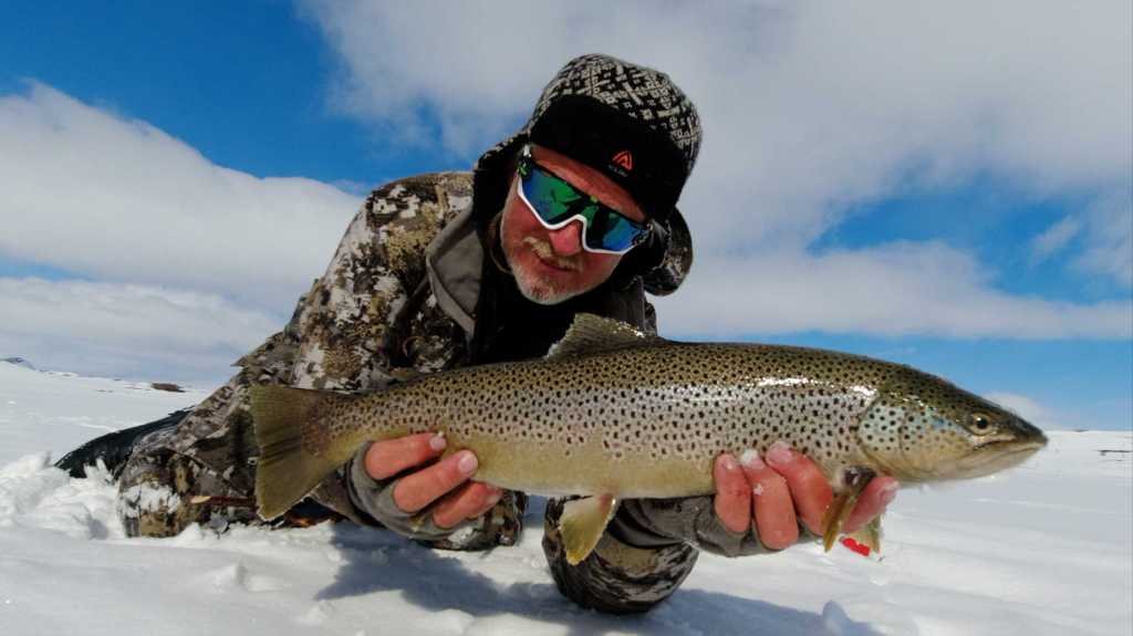 Mann ligger på isen og holder en svær ørret foran seg.