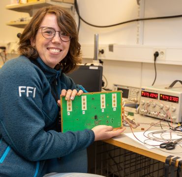 Smilende forsker i laboratorium holder opp et kretskort. I bakgrunnen ser vi masse apparater.