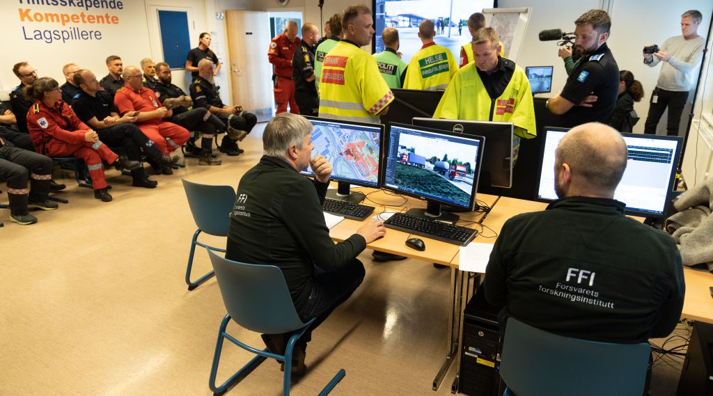 Politi, brann og ambulansepersonell står i et klasserom og diskuterer hvordan de skal løse en krisesituasjon der en tankbil full av amoniakk har kollidert og sprunget lekk. På en skjerm på veggen kan vi se en virtuell verden der krsien utspiller seg. Langs vekggen sitter kursdeltagere på to stolrader og ser på. I forgrunnen ser vi to FFI-ansatte som styrer spillet.