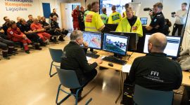 Politi, brann og ambulansepersonell står i et klasserom og diskuterer hvordan de skal løse en krisesituasjon der en tankbil full av amoniakk har kollidert og sprunget lekk. På en skjerm på veggen kan vi se en virtuell verden der krsien utspiller seg. Langs vekggen sitter kursdeltagere på to stolrader og ser på. I forgrunnen ser vi to FFI-ansatte som styrer spillet.