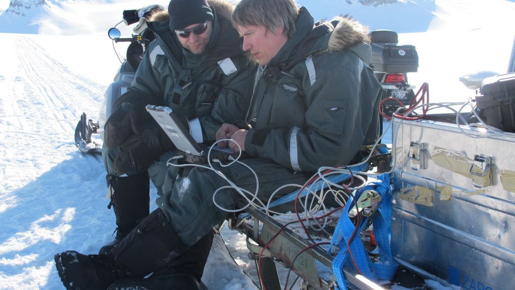 To men sitting on a snowmobile-sledge working on a laptop