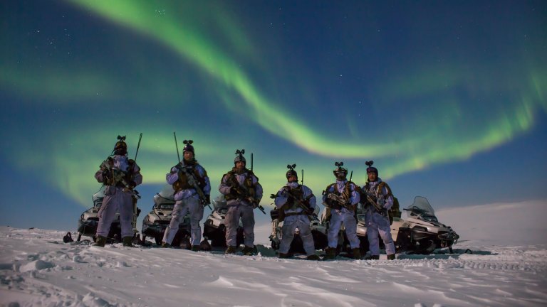 Vinterkledte soldater poserer foran snøskuter med nordlys på himmelen bak